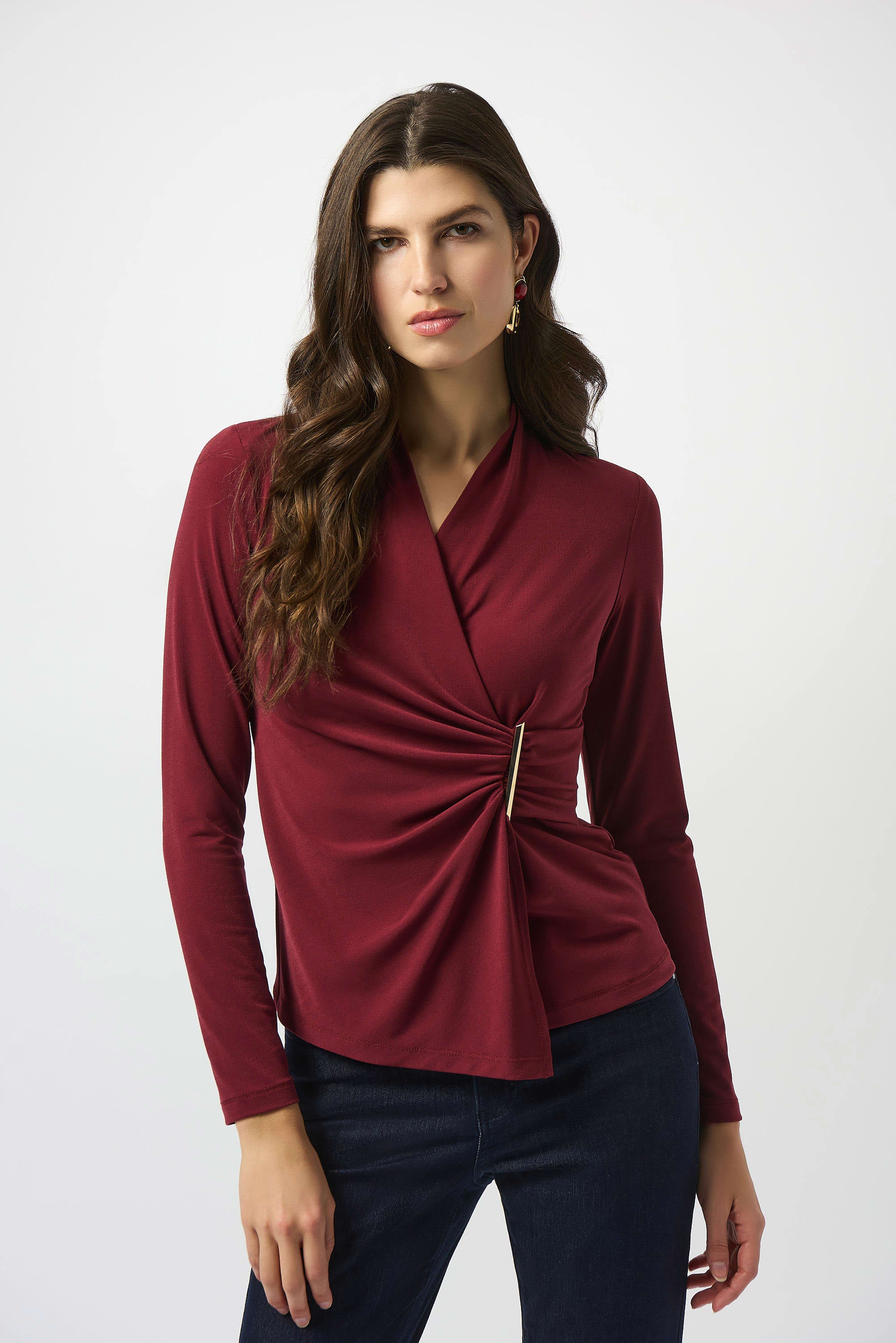A woman wearing a merlot red crepe fitted wrap top with a shawl collar and three-quarter length sleeves, featuring a pleated detail and decorative metal buttons.